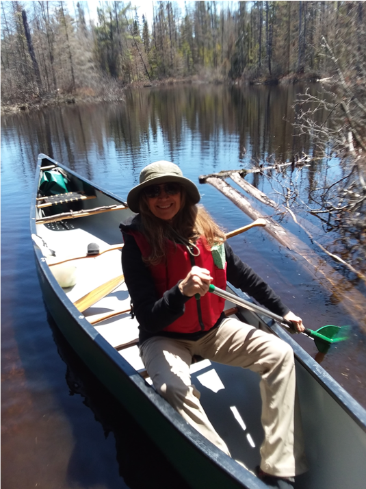 A smiling woman in a red life jacket holding an oar whilst sitting in a canoe