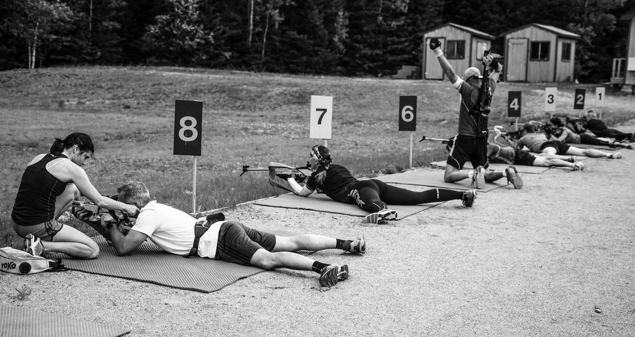 A black and white photo of a shooting fields, seven people lay in position of shooting and one person knee down on the ground and raising two hands to the sky, and all with a number sign on the right hand side, a ninth person is helping the person laying besides the number eight sign