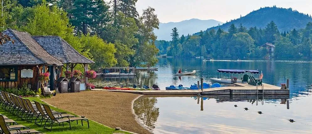 a floating deck on the shore of lake placid, two little huts next to a row of chairs on the glass land at one end of the deck, three boats on the water, forest and mountain on the opposite shore,