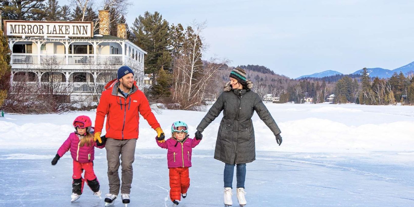 A couple holding two children's hands while skiing on snow land, a white building with the Mirror Lake Inn sign in front between the forest and the snow land in their background.