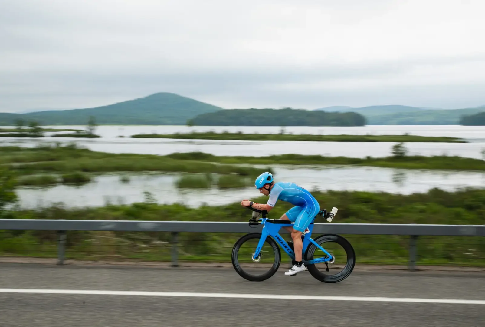 Biker in blue uniform speeds by a body of water and mountains in Tupper Lake, NY.
